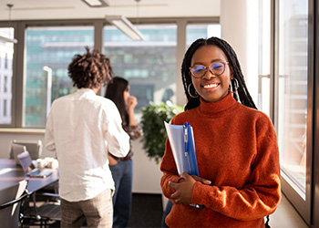 femme brune avec des tresses, porte un pull rouge-orange, des lunettes, des boucles d'oreilles, tient dans ses mains une pochette bleue (fond : salle de réunion) femme brune avec des tresses, porte un pull rouge-orange, des lunettes, des boucles d'oreilles, tient dans ses mains une pochette bleue (fond : salle de réunion)