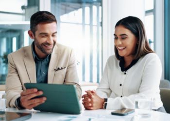 Un homme et une femme souriant, sont assis à un bureau face à la caméra et discutent en regardant une tablette tactile