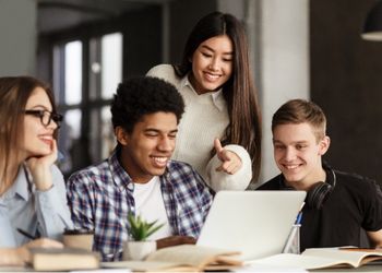 1 femme est debout derrière 1 autre femme et 2 hommes qui eux sont assis à un bureau, tous regardent un ordinateur face caméra en souriant