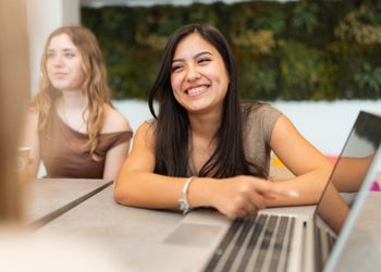 Au premier plan une étudiante souriant face caméra et pointant du doigt un ordinateur. Au second plan à gauche une autre étudiante