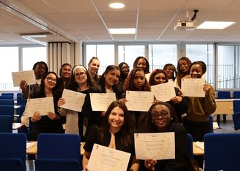 Une salle de classe avec des étudiantes debout et souriant face caméra qui tiennent et montrent face caméra leur diplome