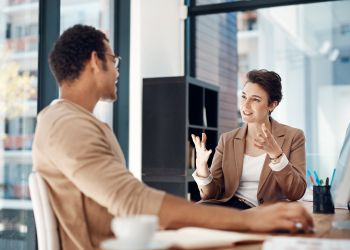 Une femme souriante face caméra en arrière plan parle avec ses mains à un homme de dos au premier plan
