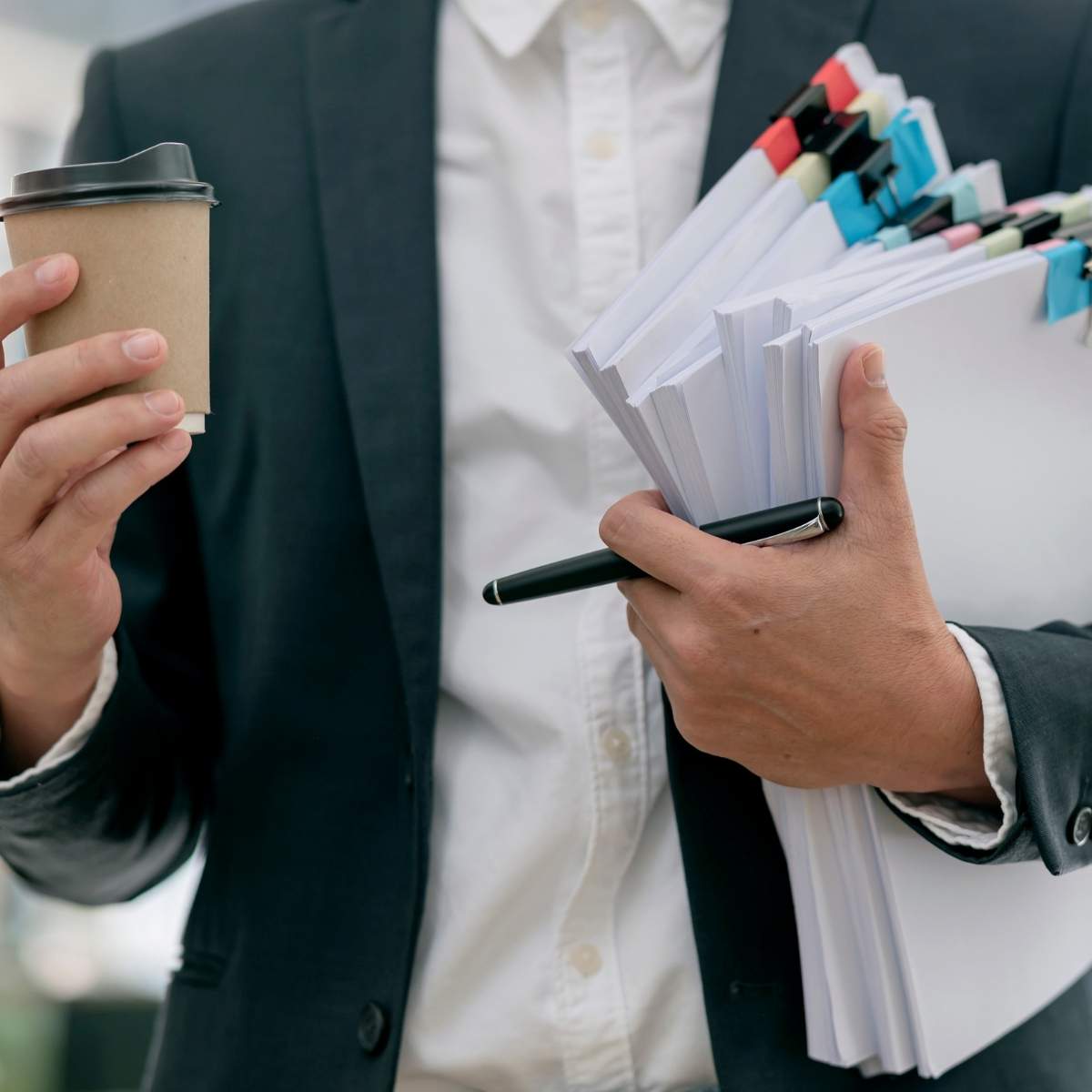 Un employé avec ses dossiers et sa boisson Une personne, en tailleur et chemise, porte des dossiers et un stylo de la main gauche puis un gobelet de la main droite