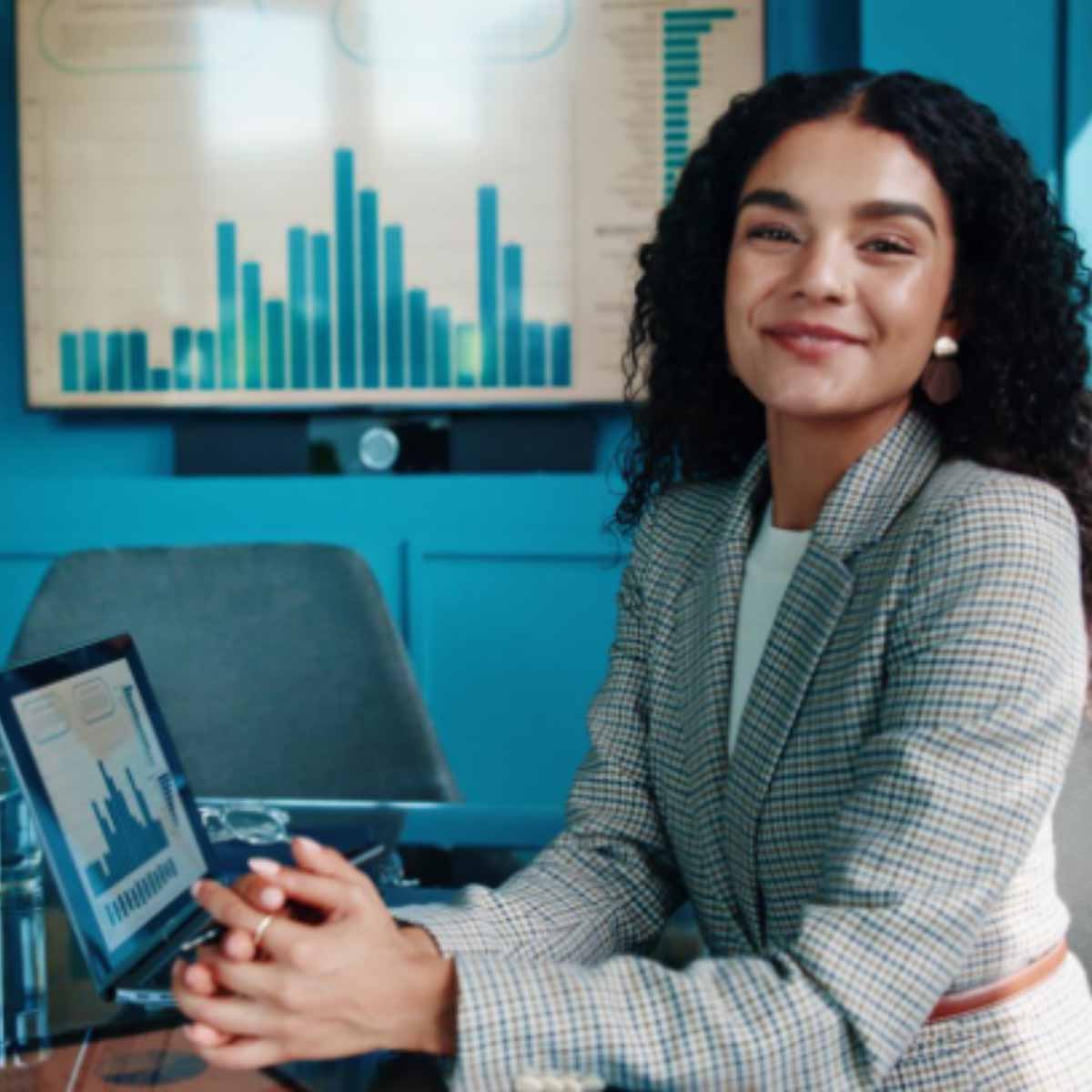 Employé RH Une femme souriante regardant face caméra assise à un bureau avec dessus un ordinateur et sur le mur du fond un histogramme projeté