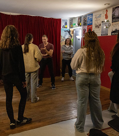 Professeur parle avec une étudiante, des étudiants sont en cercle autour d'eux dans un théâtre rouge