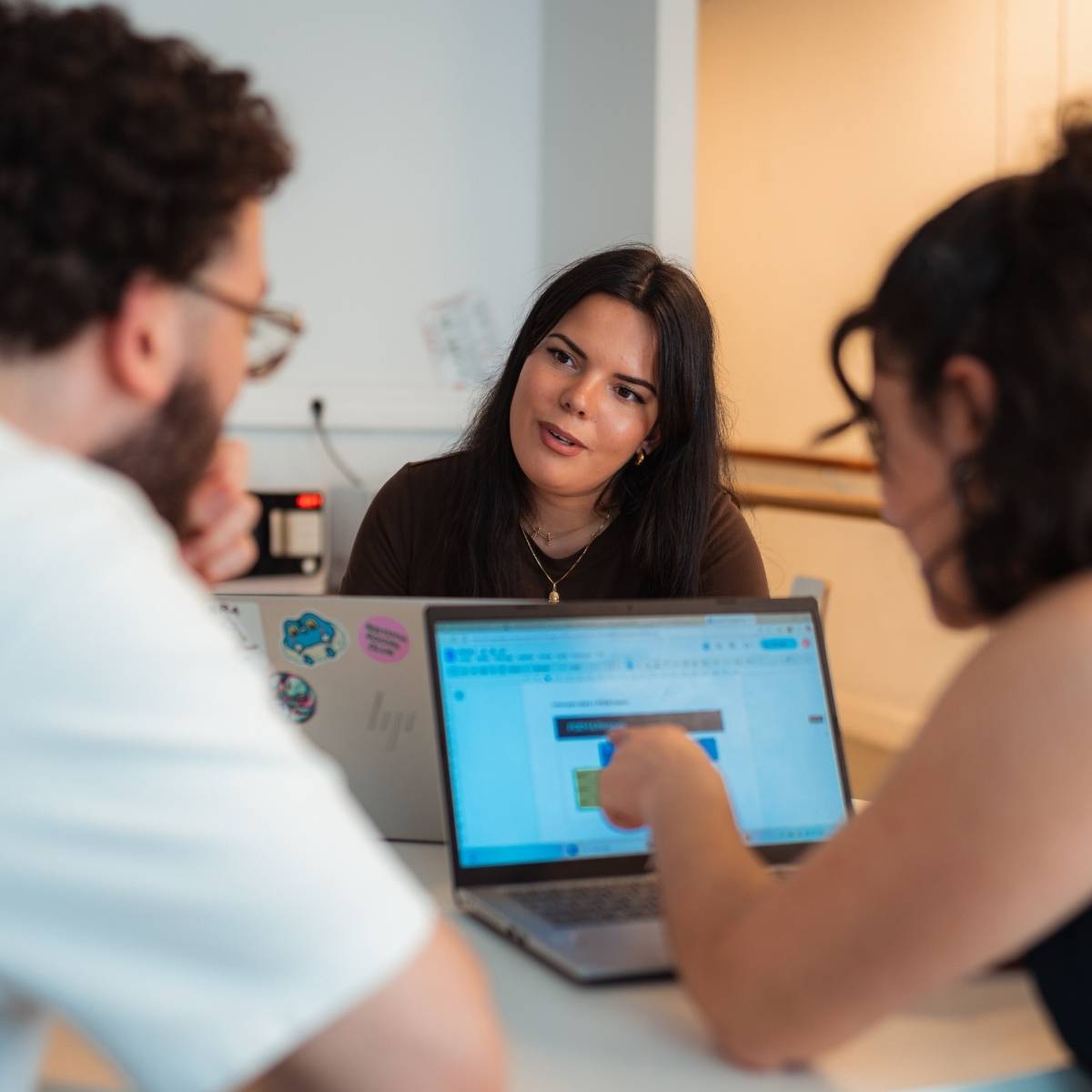 Une femme cheveux bruns longs assise à un bureau face caméra et face à un homme cheveux bruns courts et bouclés et une femme cheveux bruns courts et bouclés sont eux dos à la caméra. Ils discutent avec des ordinateurs face à eux 