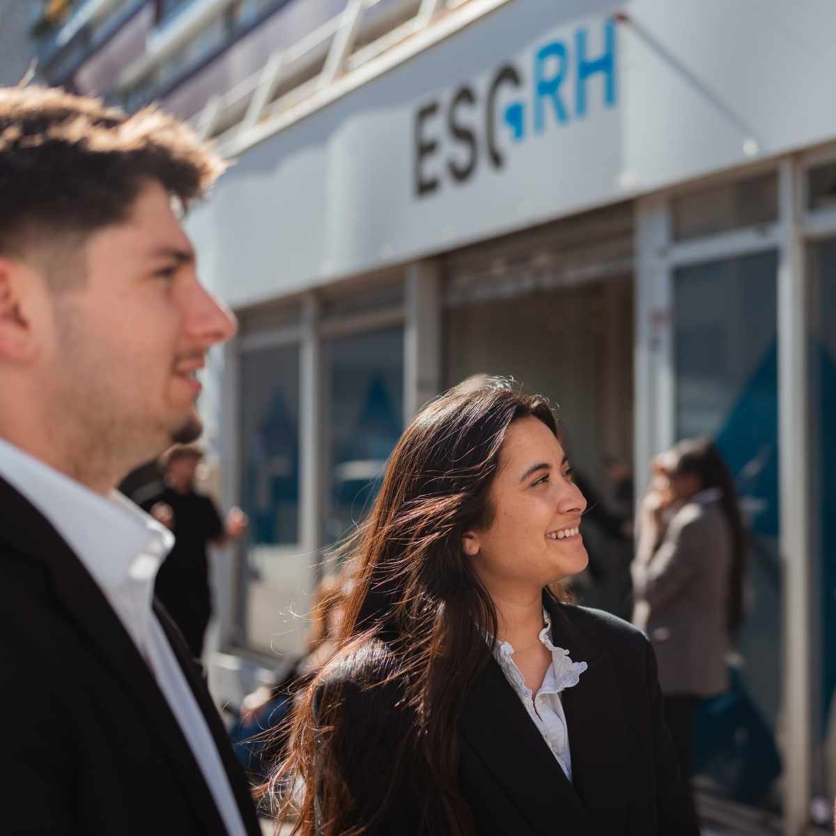 En extérieur devant le campus de l'ESGRH avec au premier plan un étudiant de côté qui sourit et au second plan une étudiante de côté et qui sourit aussi