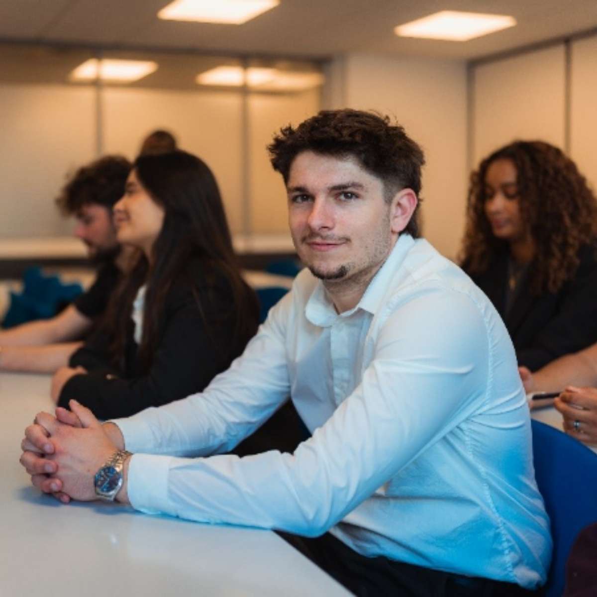 Salle de classe avec au premier plan un étudiant assis à sa table souriant face caméra et au second plan des étudiants