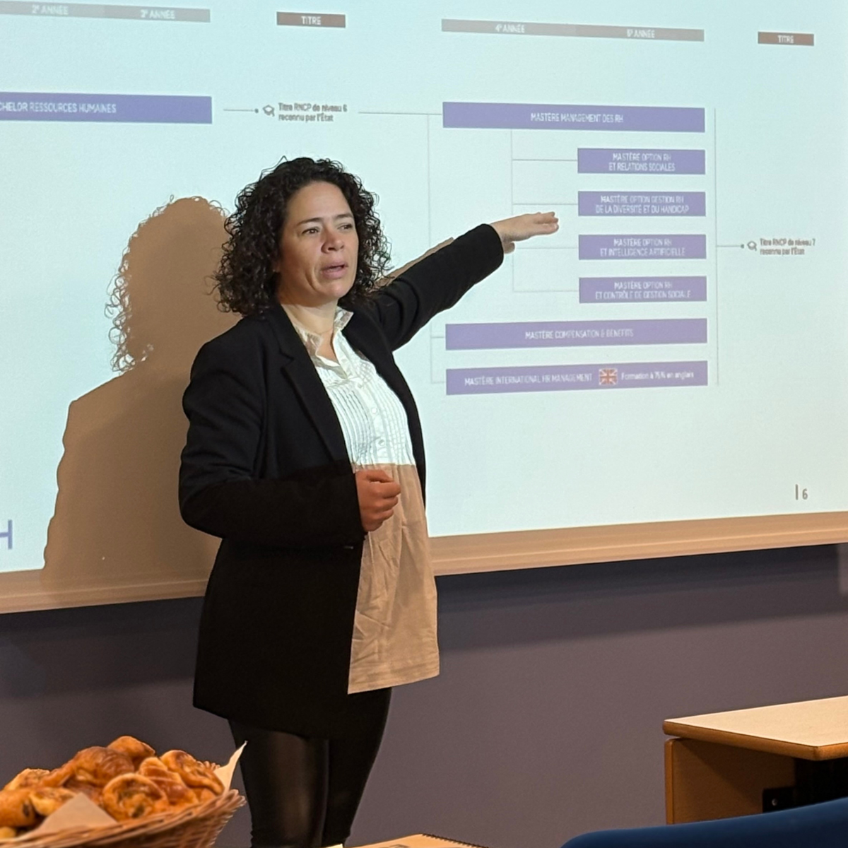 Femme costume noir et cheveux noirs, courts et bouclés, donne une conférence aux tuteurs dans une salle avec des murs bleus