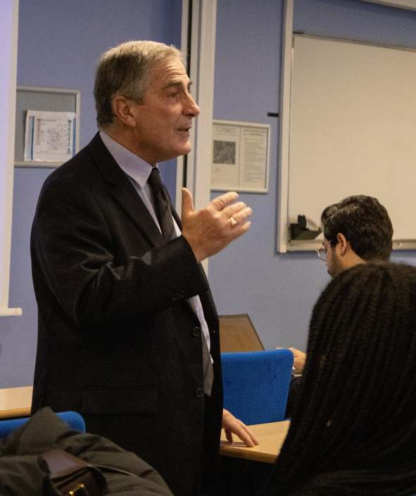 Homme costume noir et cheveux blancs, donne une conférence à étudiants dans une salle avec des murs bleus assis sur chaises bleues